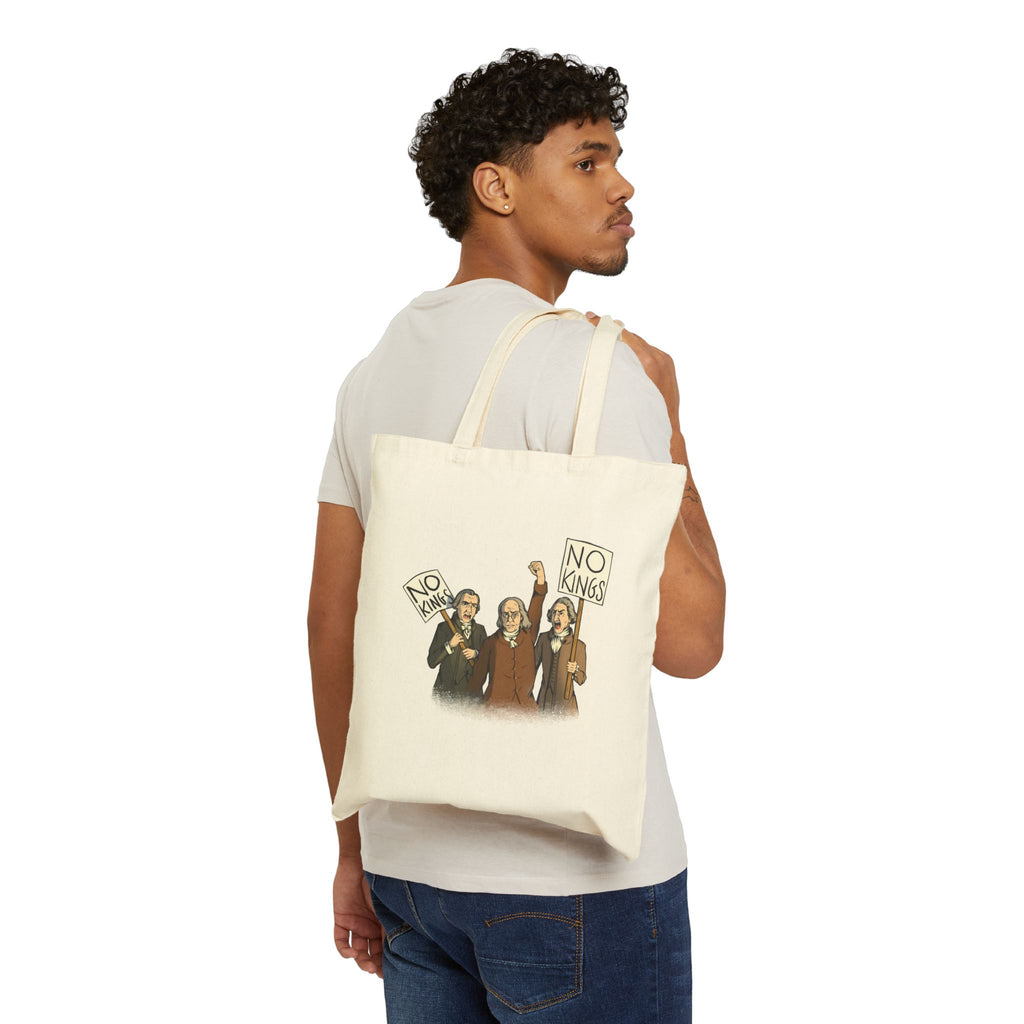 A man wearing a white t-shirt and blue jeans is shown from the back, carrying a cream-colored canvas tote bag over his shoulder. The tote bag features a printed graphic of three Disappointed American Founders protesting and shouting, with two of the men holding signs that read NO KINGS