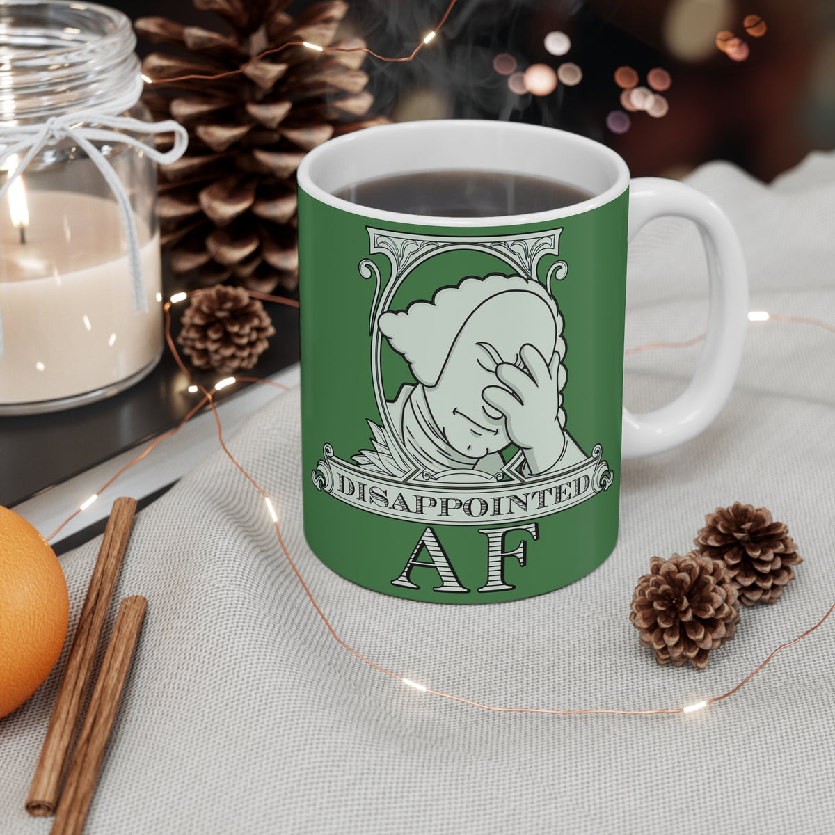 A white ceramic mug featuring a green logo for Disappointed AF. The logo is white and framed with scrolls, similar to that of a dollar bill. George Washington's bust is in the center, looks disappointed, is leaning his head forward, and has his head in his hand with an exasperated look on his face. Below the figure it says Disappointed AF. The mug is sitting on a table surrounded by cozy decor such as two pinecones, cinnamon sticks, and a lit candle.