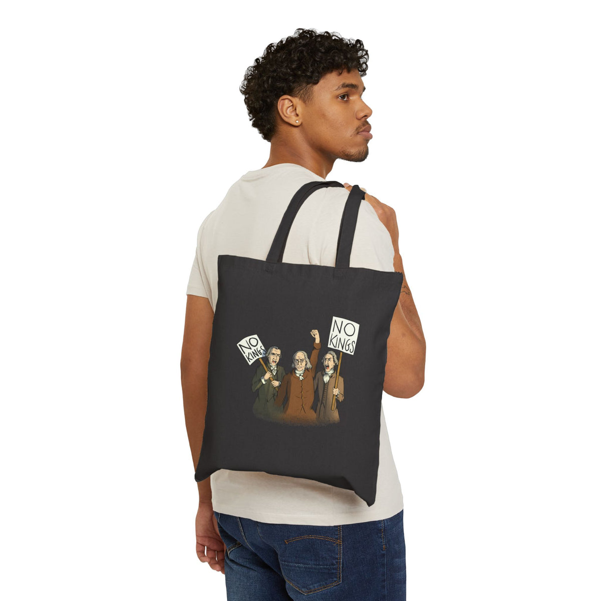 A man wearing a white t-shirt and blue jeans is shown from the back, carrying a black canvas tote bag with two straps over his shoulder. The tote bag features a printed graphic of three Disappointed American Founders protesting and shouting, with two of the men holding signs that read NO KINGS