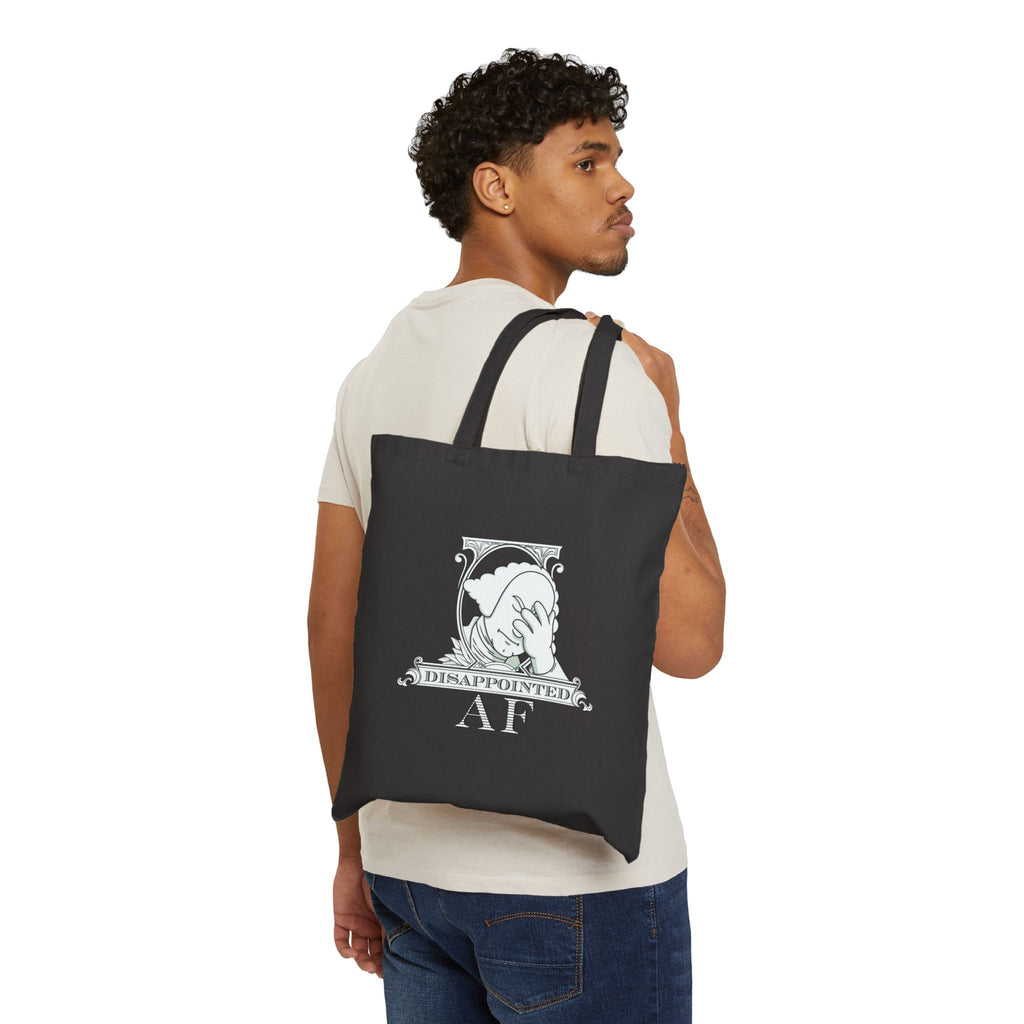 A black man wearing jeans and a white tshirt is carrying a black tote bag with a printed design on a white background. The bag has the logo for Disappointed AF printed on it. The logo is framed with scrolls, similar to that of a dollar bill, with George Washington in the center. George Washington looks disappointed and has his head in his hand. below the logo it says Disappointed AF