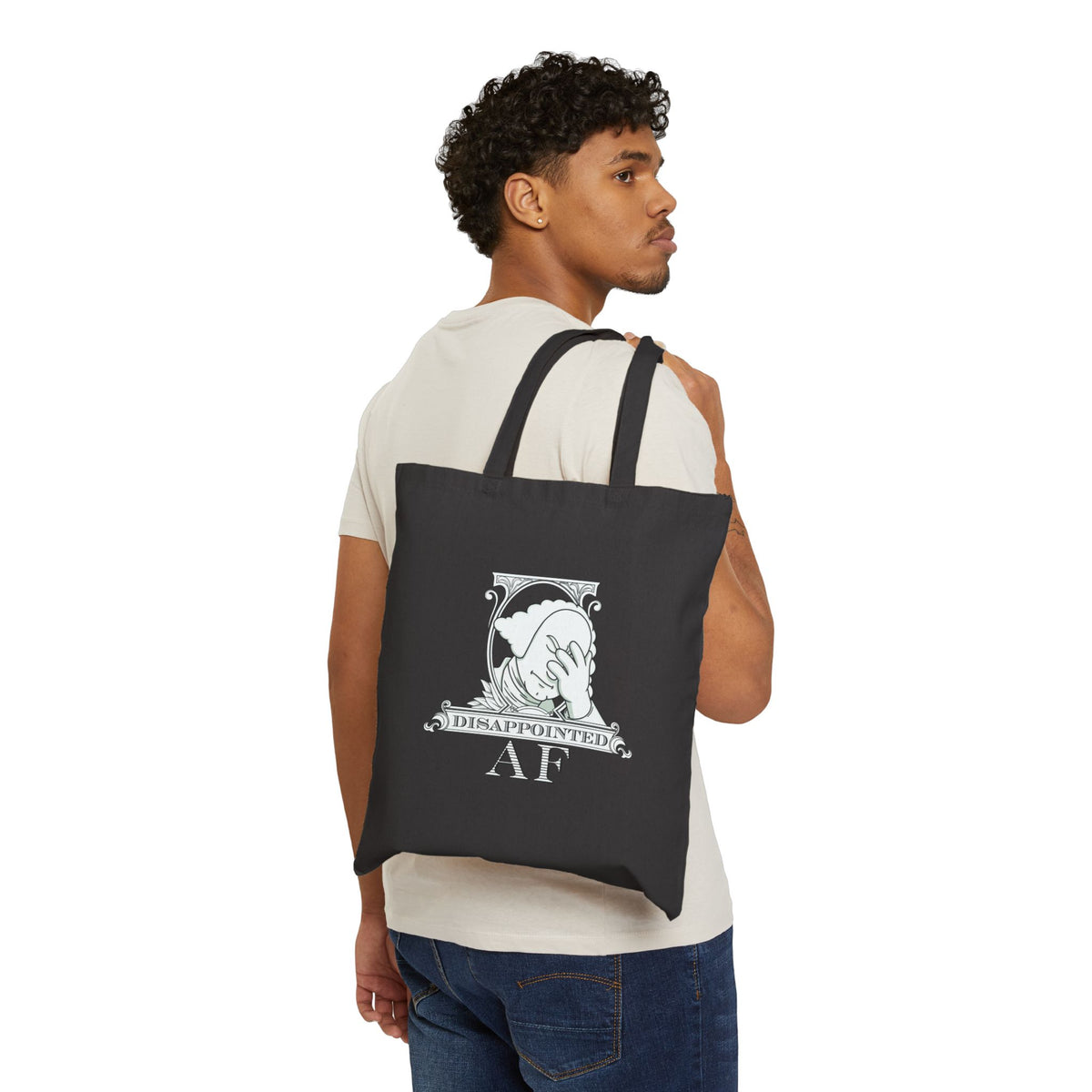 A black man wearing jeans and a white tshirt is carrying a black tote bag with a printed design on a white background. The bag has the logo for Disappointed AF printed on it. The logo is framed with scrolls, similar to that of a dollar bill, with George Washington in the center. George Washington looks disappointed and has his head in his hand. below the logo it says Disappointed AF